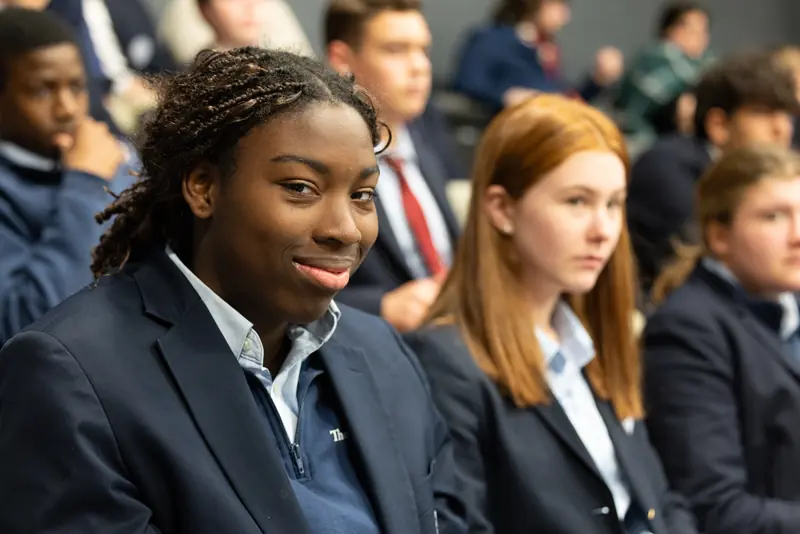 Students in navy blazers sit together in the audience at Assembly, one smiling at the camera
