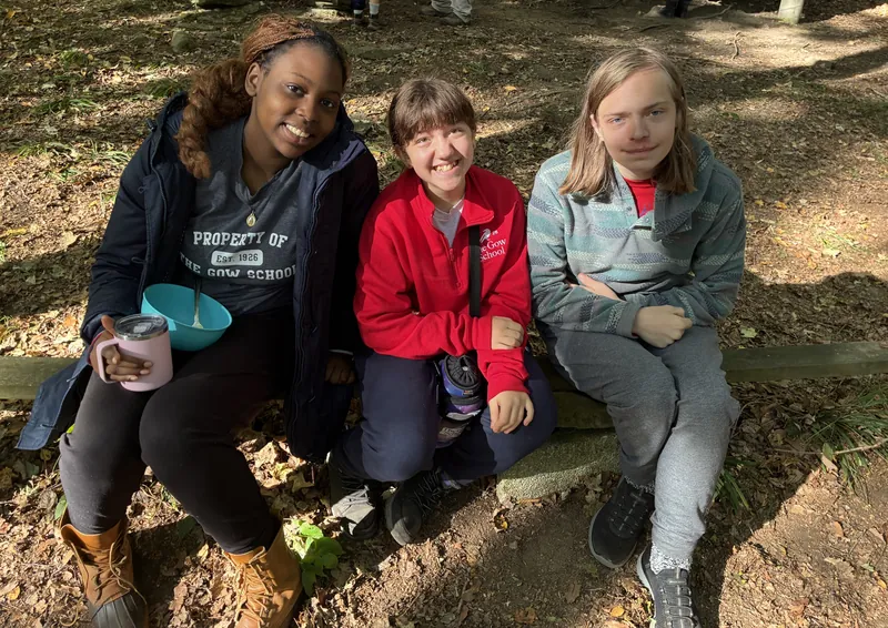 Three Gow students smile together while sitting on a log in the woods