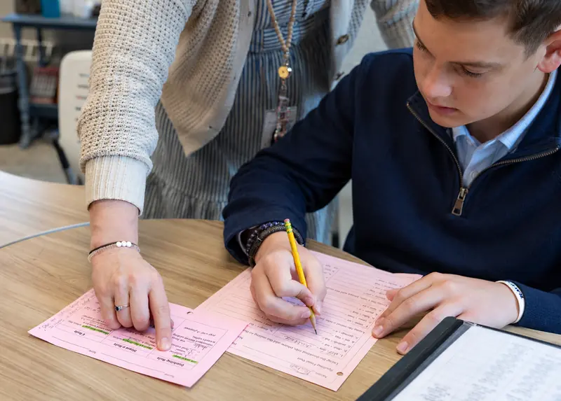 A teacher points to a line on a pink worksheet as a student in a navy sweater writes his answer