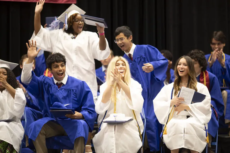 Graduates in blue and white caps and gowns cheering, clapping and waving with their diplomas at Commencement
