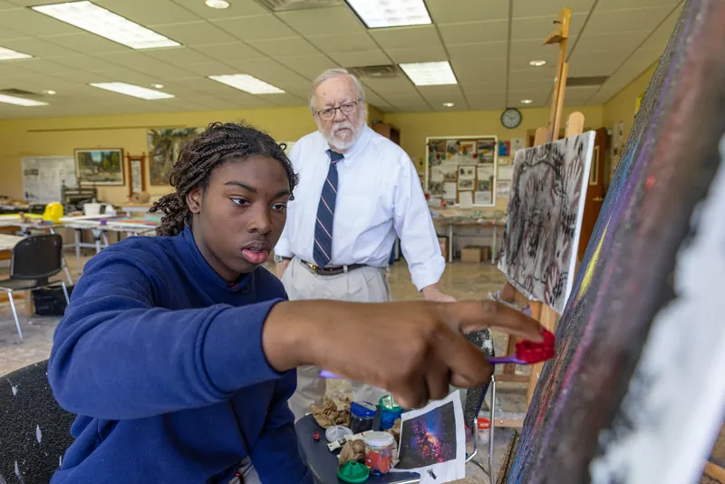 A student painting at an easel in the Reid Art Center as her art teacher looks on