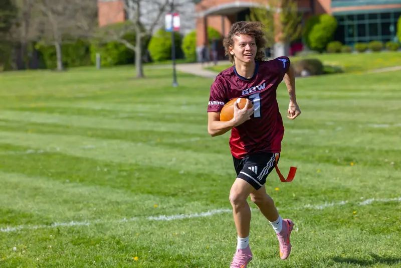 A student in a crimson Gow jersey runs downfield with a football during a flag football game on the campus lawn