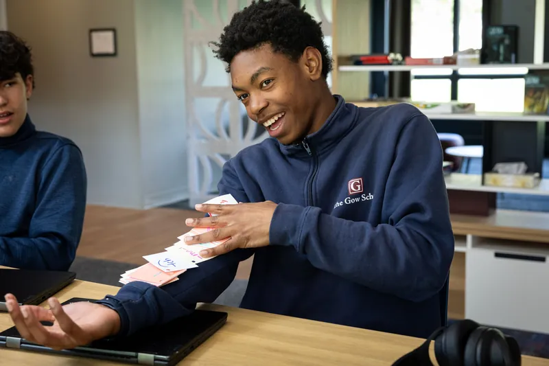 A laughing student in a navy Gow quarter-zip fans out a stack of handwritten Gow Method phonics cards along one arm, reviewing them with a classmate