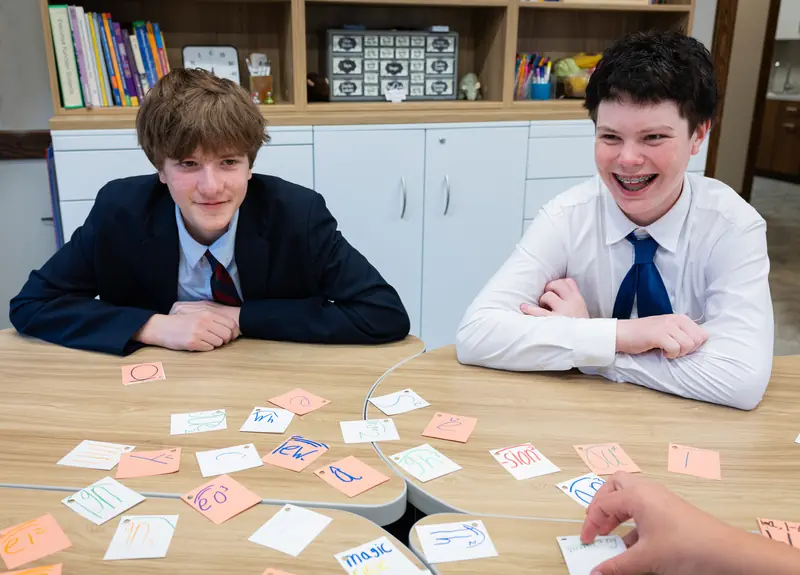 Two students in shirts and ties lean on a classroom table and laugh as they review Gow Method phonics cards spread out in front of them