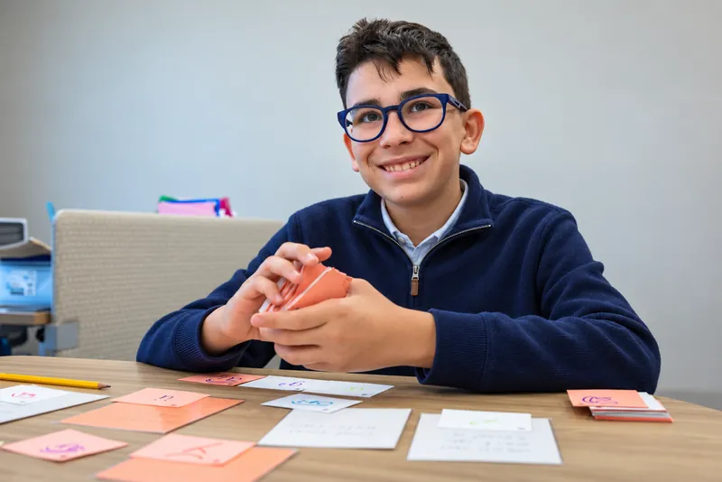 A smiling middle school student in glasses reviews a deck of Gow Method phonics cards, with more cards spread across the table