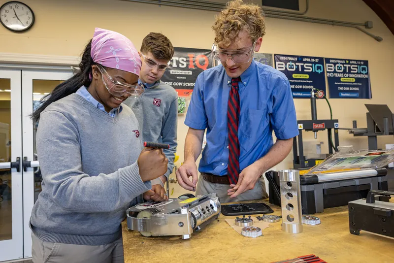 Three students assembling a combat robot at a workbench in front of BotsIQ banners