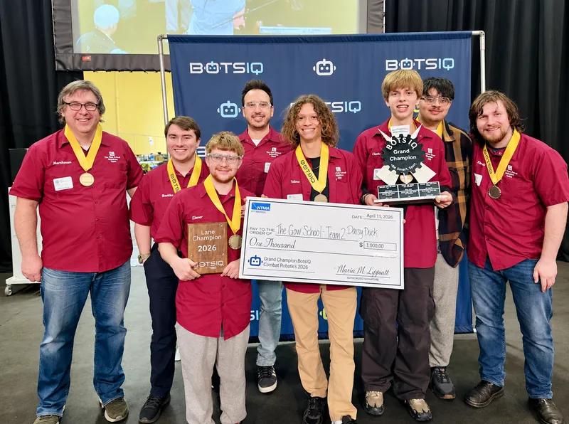 The Gow robotics team and coaches in crimson shirts and medals, holding the Grand Champion trophy and a prize check at BotsIQ in 2026