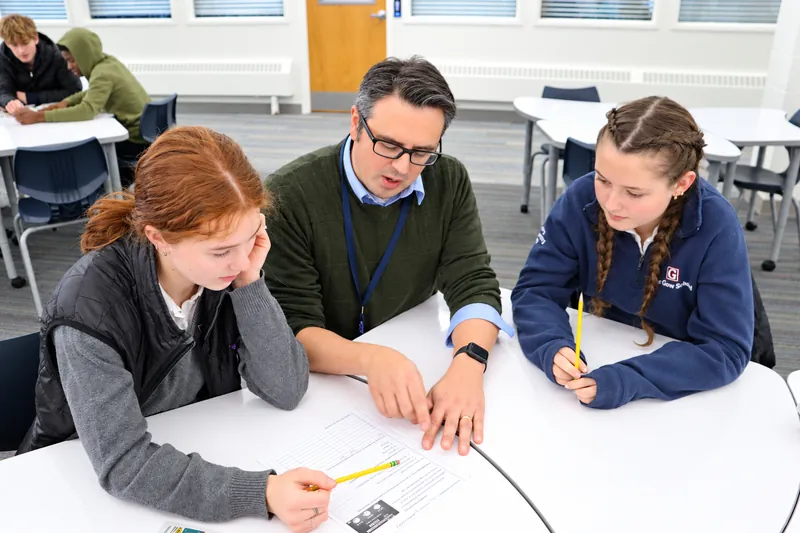 A Gow teacher points to a worksheet as two students follow along at a round table in a bright classroom