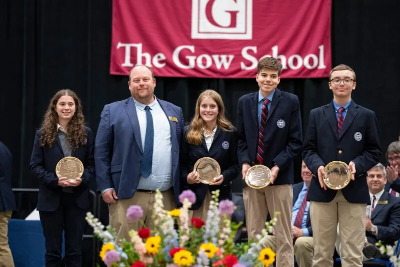 Four students in navy Gow blazers hold silver award plates on stage beside a faculty member, beneath a crimson The Gow School banner