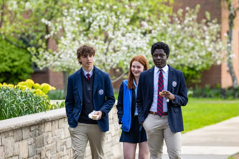 Three Gow students in navy blazers walk along a stone wall past spring flowers and a blossoming tree
