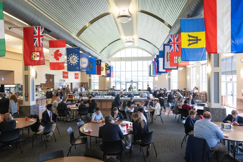 Students and faculty eat at round tables in the Weston Dining Hall beneath flags from students' home states and countries