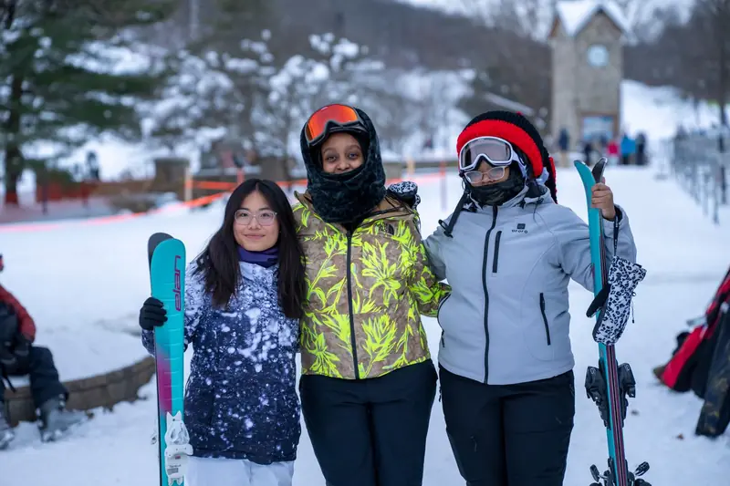 Three students in ski jackets and goggles smiling arm in arm, holding their skis at the bottom of a snowy ski hill