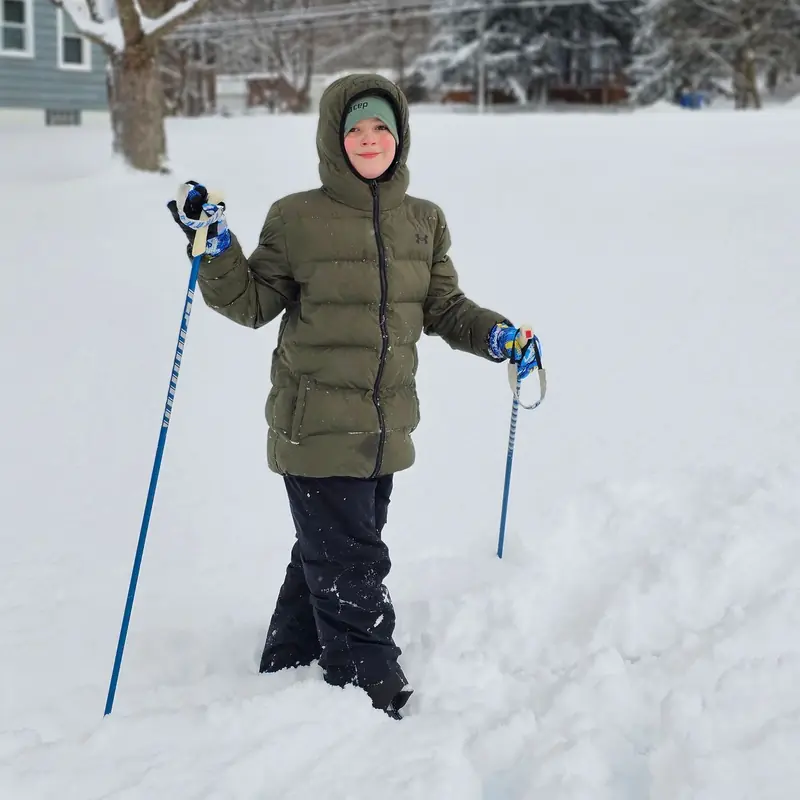 A smiling young skier in a hooded green winter jacket standing knee-deep in fresh snow with ski poles